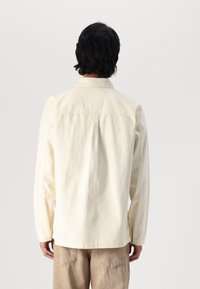 Light cream long-sleeve shirt with a collar, stitched detailing, and a back pleat. Paired with beige pants. Smooth cotton texture.