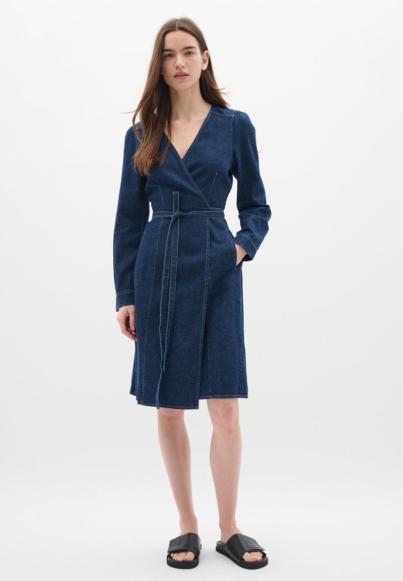 Denim omslagsklänning i mörkblått med långa ärmar, sidfickor och ett tygbälte. Har kontrasterande sömmar och en knälång skärning.