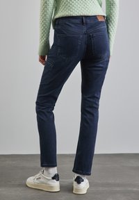 Jean en denim bleu foncé à coupe slim, avec deux poches arrière et une poche latérale. Porté avec des baskets blanches et un pull vert texturé.