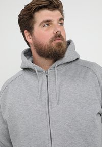 Sudadera gris con cremallera hecha de una tela suave, que cuenta con capucha, cordones y costuras de contraste a lo largo de los hombros y los lados.