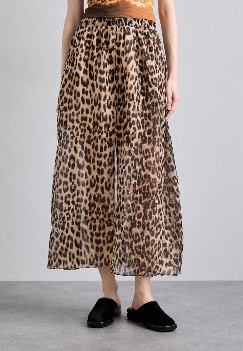 Femme portant une jupe midi imprimé léopard avec une taille élastique et des chaussures en fourrure noires à enfiler, debout sur un sol gris.