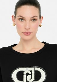 Must mustusega tipp, millel on valge pärli ja kivist logodisain. Modell kannab kuldseid südamekujuliselt kõrvakeesid, millel on dekoratiivne detail.
