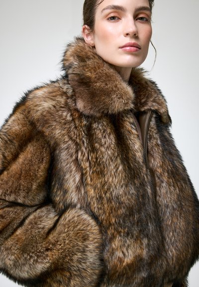 Jeune femme portant un épais manteau en fourrure marron et noir avec un grand col, posant devant un fond clair uni.