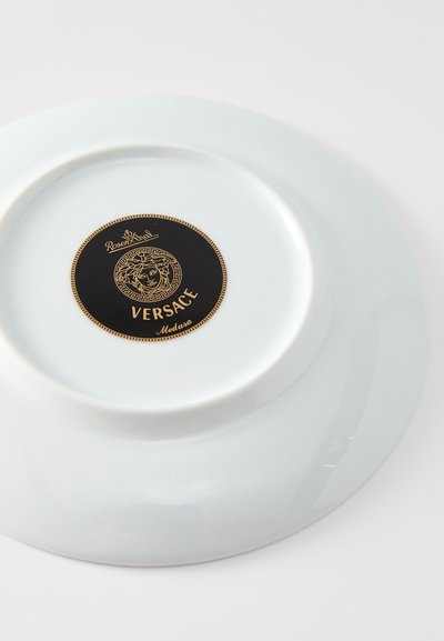 Κυκλικό λευκό πορσελάνινο πιάτο, με μαύρη και χρυσή διακοσμητική ετικέτα με λογότυπο Μέδουσας. Λείο, γυαλιστερό στέφανο με ανυψωμένη στεφάνη.