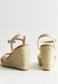 Sandales à talon compensé beige avec un talon en jute tissé, une sangle pour la cheville et un design à bout ouvert. Texture en cuir lisse avec un accent en bois naturel.