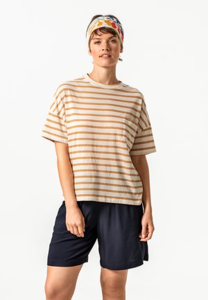 Woman wearing a beige and white striped t-shirt, navy shorts, and a colorful floral headband, standing against a white background.