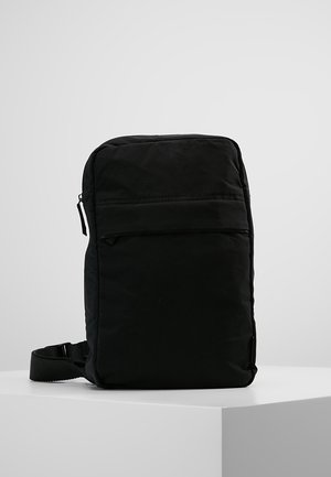 Bolso bandolera compacto y rectangular de color negro, con bolsillo frontal con cremallera y correa ajustable, exhibido sobre una superficie blanca contra un fondo gris claro.