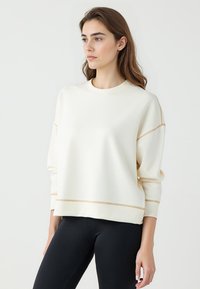 Kremowy sweatshirt z okrągłym dekoltem, opadłymi ramionami i kontrastowymi złotymi ściegami wzdłuż dołu oraz rękawów.