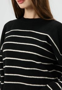 Pull noir avec des rayures horizontales blanches, col et poignets côtelés, texture douce, coupe décontractée, mettant en valeur un design moderne et décontracté.