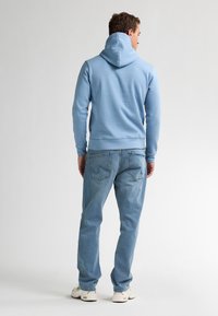 Sudadera azul claro con capucha ajustable, textura suave, combinada con vaqueros azul claro y zapatillas blancas, vista desde la parte trasera.