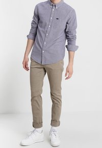 Camisa de botón de chambray en azul claro con un bolsillo, combinada con pantalones slim-fit en caqui y zapatillas blancas, con los puños enrollados.