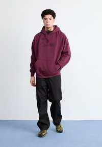 Felpa bordeaux con tasca frontale e logo ricamato, abbinata a pantaloni neri e scarpe mimetiche verdi, in piedi su un pavimento blu.