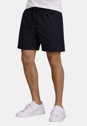 Man wearing black drawstring shorts and white sneakers standing against a plain background.