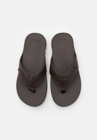 Bruine flip-flops met een getextureerde rubberen zool, leren banden en een metalen accent. De banden hebben een zigzagsteek en geperforeerde details.