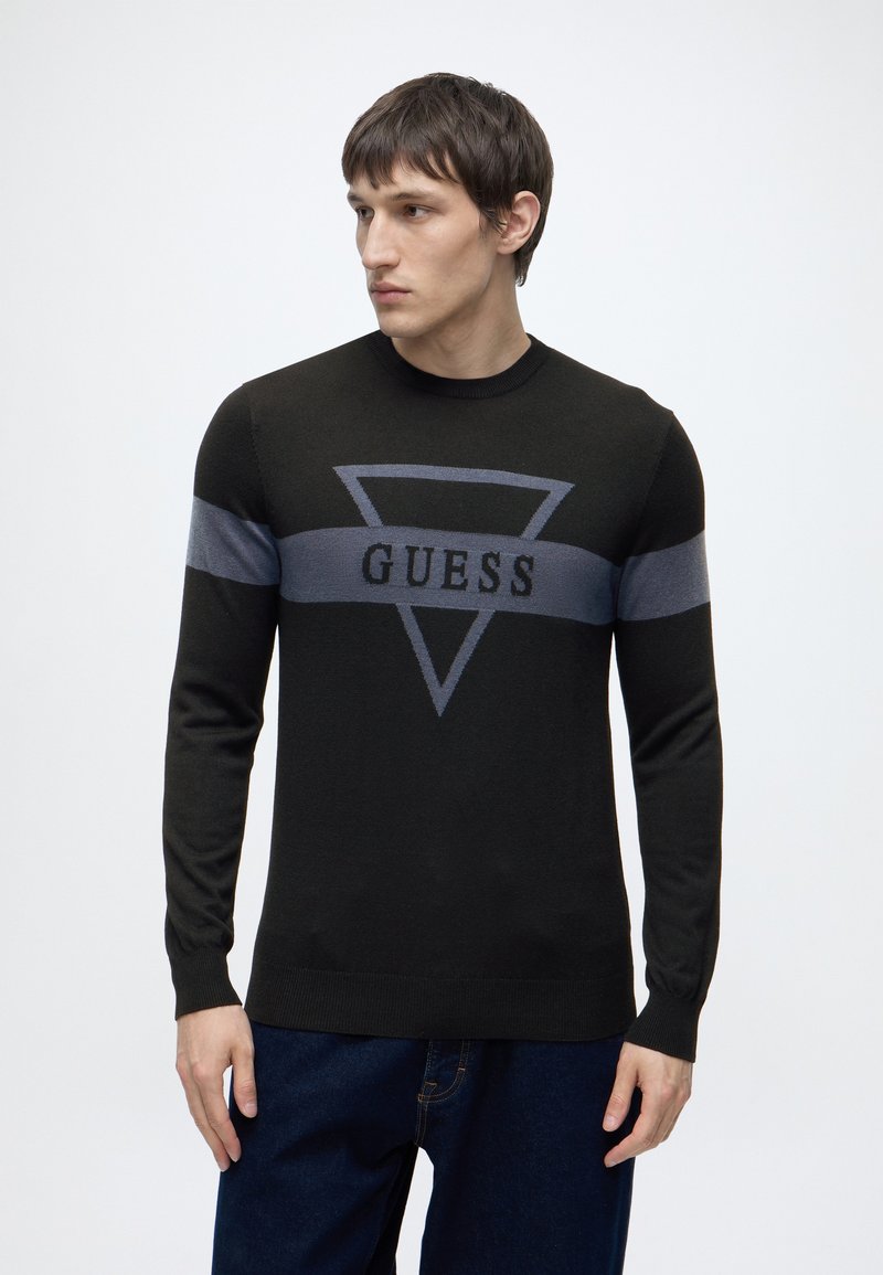 Jeune homme portant un pull noir avec une bande horizontale grise et un motif triangulaire avec le mot « GUESS » sur la poitrine.