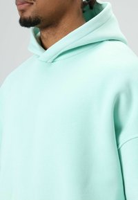 Sweat à capuche vert menthe avec une texture douce, doté d'une capuche à cordon de serrage et d'une conception sans coutures aux épaules. Style simple et décontracté.