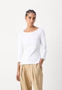 Camiseta blanca de manga larga con un escote redondeado, hecha de una suave tela de algodón; combinada con pantalones de cintura alta de color beige claro con pliegues.