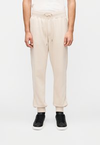 Μπεζ φλις φόρμα με ελαστική ζώνη μέσης, κορδόνι, πλαϊνές τσέπες και ριμπ μανσέτες. Συνδυασμένο με μαύρα αθλητικά παπούτσια.