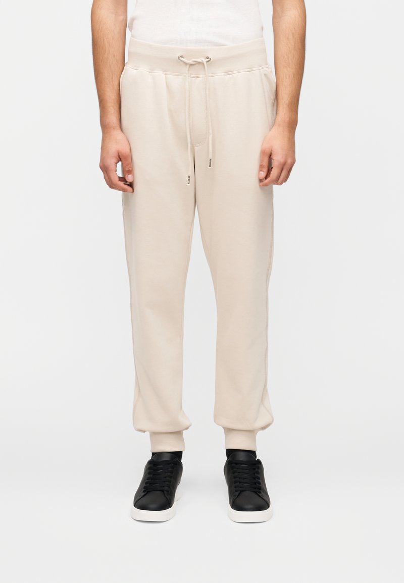 Μπεζ φλις φόρμα με ελαστική ζώνη μέσης, κορδόνι, πλαϊνές τσέπες και ριμπ μανσέτες. Συνδυασμένο με μαύρα αθλητικά παπούτσια.