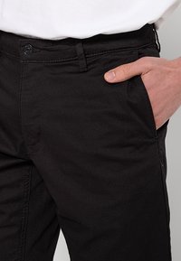 La main droite repose dans la poche avant du pantalon noir porté avec une chemise blanche.
