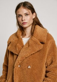 Manteau en shearling brun croisé avec grands revers, surface texturée et deux boutons ronds. Porté sur un haut blanc.