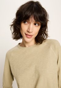 Junge Frau mit kurzen, dunklen, welligen Haaren, die ein hellbeiges Langarmoberteil trägt und leicht nach links schaut, mit einem sanften Lächeln.