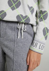Šedý svetr se srdíčkovými vzory a viditelnými neonově zelenými liniemi přes šedé tepláky. Tepláky mají stahovací šňůrku a boční kapsy.