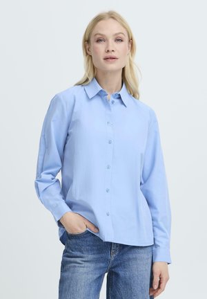 Donna bionda con camicia azzurro chiaro abbottonata e jeans blu, in piedi con una mano in tasca contro uno sfondo semplice.