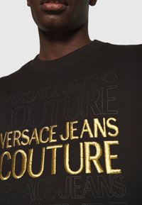 Černá mikina s velkým zlatým výšivkou "VERSACE JEANS COUTURE" a jemným černým textem, vystihující texturovanou tkaninu a design s kulatým výstřihem.