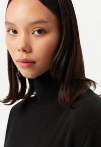 Svart stickad polotröja med ribbad hals. Har slät textur och avslappnad passform. Modellen har långt mörkt hår och neutral makeup.