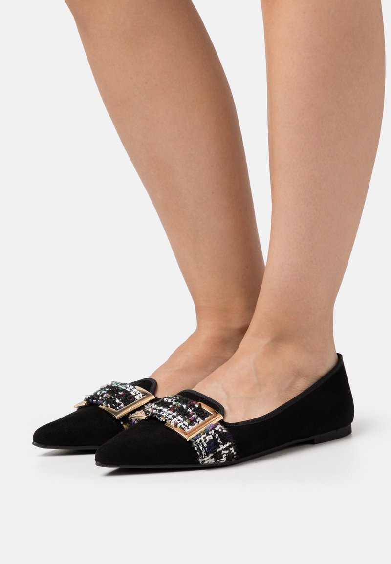 Pieds chaussés de ballerines noires à bouts pointus, ornées de sangles en tweed multicolore et de boucles dorées, sur un fond clair uni.