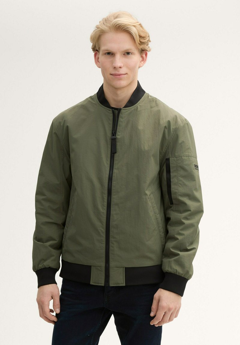 Olivgrön bomberjacka, tillverkad av lättviktsmaterial, har en framsida med dragkedja, svarta ribbade muddar och en sidoficka med logodetalj.