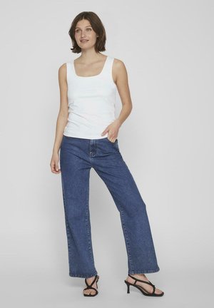 Weißes Tanktop mit quadratischem Ausschnitt, kombiniert mit weiten blauen Jeans. Das Model trägt schwarze Riemchen-Heels. Glatte Stoffqualität und lässige Passform.