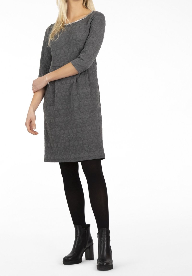 Femme portant une robe grise texturée longueur genou, à manches trois-quarts, collants noirs et bottines noires à talons.