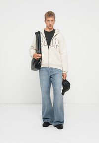 Sudadera crema con cremallera y logotipo "GAP", camiseta negra, vaqueros anchos de mezclilla azul, bolso negro, zapatos negros. La modelo está de pie con las manos relajadas.