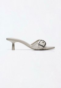 Mule beige à bout ouvert avec un petit talon bas et une grande boucle argentée sur la bride avant.