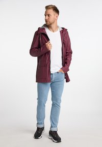 Bordeauxfarvet vandtæt jakke med grå hætte, udstyret med trykknapper, sidelommer og en let tætsiddende pasform. Båret over en hvid skjorte og blå jeans.