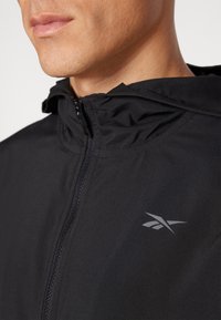Chaqueta negra con cremallera y capucha, hecha de tela suave. Presenta un logo gris sutil en el lado frontal y un cuello alto con cremallera.
