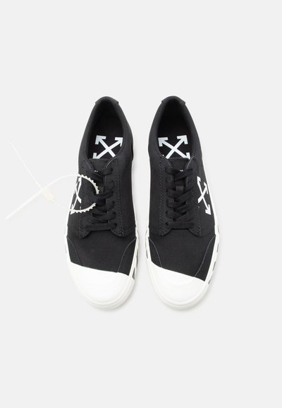 Ζευγάρι μαύρα αθλητικά παπούτσια χαμηλού ύψους με άσπρες λαστιχένιες μύτες, λευκά λογότυπα βέλους σε σχήμα σταυρού στις εσωτερικές σόλες και τις πλευρές, και κυκλικό ταμπελάκι προσκολλημένο στο αριστερό παπούτσι.