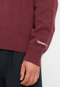 Suéter de punto burdeos con un acabado texturizado, dobladillo y puños acanalados, que presenta texto bordado en la manga. Combinado con pantalones a rayas.