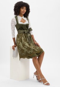 Frau sitzt auf einem weißen Block und trägt ein olivgrünes floral-lace Kleid mit Satin-Schleife, eine weiße Spitzenbluse, silberne Absätze und hält eine kleine Clutch.