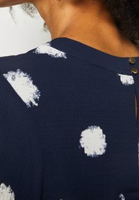 Hombros y parte superior de la espalda de una persona que lleva una tela azul marino con manchas blancas que parecen pintura y dos botones de bronce cerca del escote.