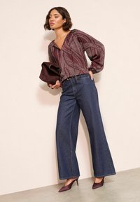 Bordeauxfarbener Blusen mit Muster, langen Ärmeln und V-Ausschnitt, kombiniert mit weit geschnittenen dunkelblauen Jeans. Sie hält eine burgunderfarbene Clutch.