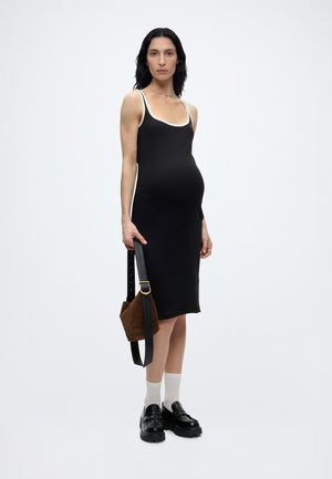 Έγκυος γυναίκα στέκεται, φοράει μαύρο αμάνικο φόρεμα με λευκή επένδυση, λευκές κάλτσες, μαύρα χοντρά παπούτσια και κρατά καφέ τσάντα.