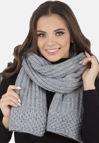 Écharpe en tricot gris avec des motifs côtelés et texturés, fabriquée en fil doux. Présente un tissage épais avec des extrémités frangées et une apparence chaleureuse.