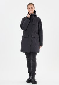 Svart, medellång jacka med huva, flera framfickor och dragkedja. Bärs över mörkgrå jeans och svarta ankelboots. Slät textur.