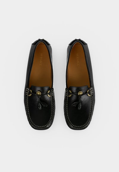 Paire de mocassins en cuir noir avec emblème Méduse doré et détail pompon, vus de dessus sur fond blanc.