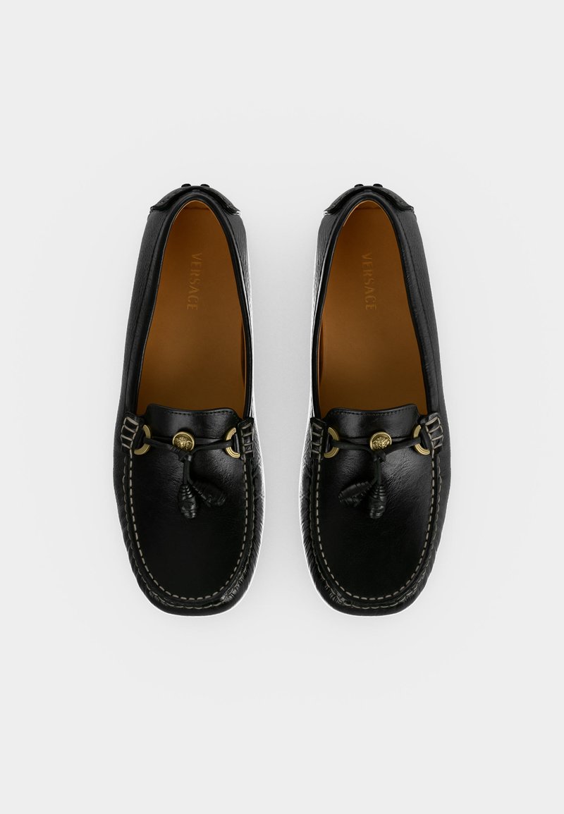 Paar schwarze Leder-Loafer mit goldfarbenem Medusa-Emblem und Quasten-Detail, von oben auf weißem Hintergrund betrachtet.
