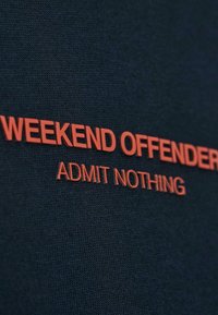 Červený text "WEEKEND OFFENDER" a "ADMIT NOTHING" na tmavom textúrovanom pozadí, pravdepodobne súčasť oblečenia.