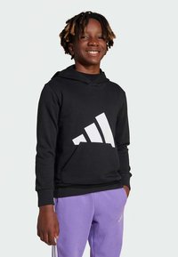 Sweatshirt preta com capuz, com logo branco da Adidas, punhos e bainha canelados. Usada com calças de fato de treino lilás claro. Textura de tecido lisa.